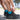 A person ties the laces of black running shoes on a sunny day. They wear bright blue socks, standing on a paved path with blurred greenery in the background.